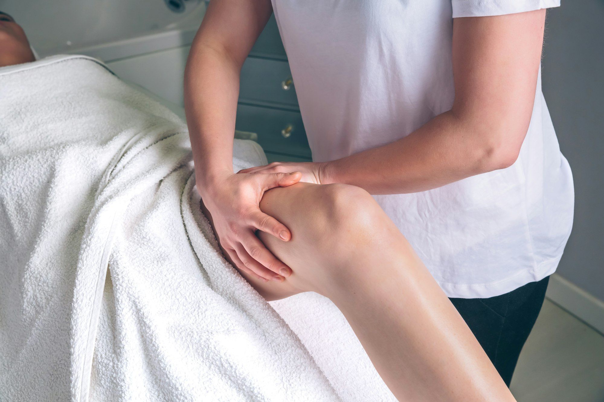 Pilates Canal Madrid Una persona recibe un masaje en las piernas mientras está acostada, cubierta con una toalla blanca. Las manos del masajista se centran en la zona de las rodillas, realizando un drenaje linfático manual en Madrid. El masaje se lleva a cabo en un entorno profesional.