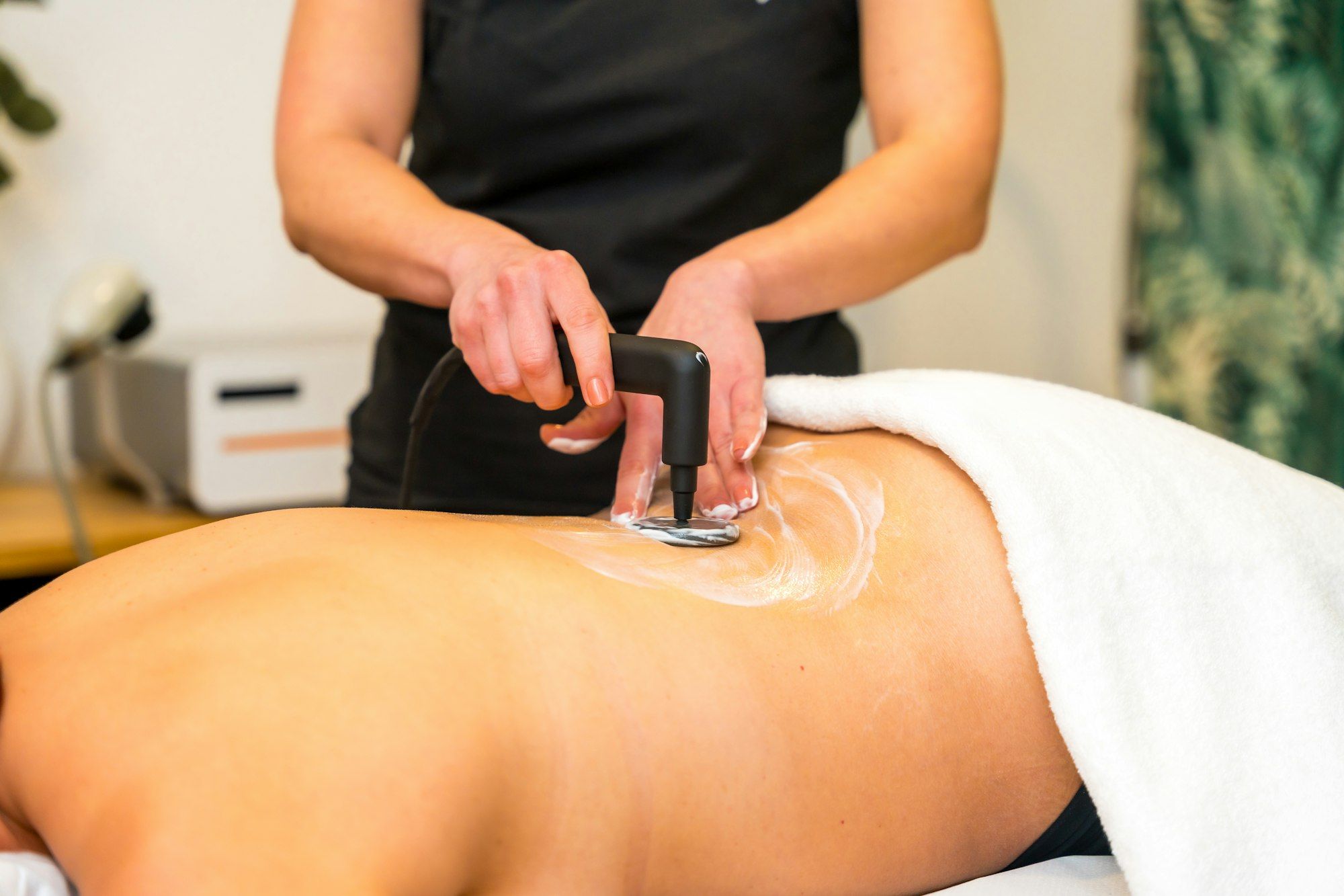 Pilates Canal Madrid Una persona recibe un tratamiento en la espalda con un dispositivo terapéutico portátil en un entorno de spa, centrado en la fisioterapia dermatofuncional. El terapeuta presiona el dispositivo contra la espalda de la persona, cubierto con un gel transparente, mientras una toalla blanca protege la parte inferior del cuerpo.