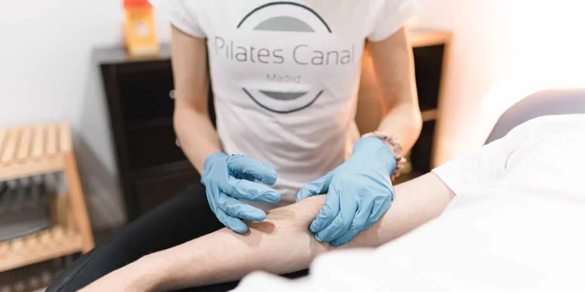 Pilates Canal Madrid Una persona con guantes azules y una camiseta de Pilates Canal realiza una punción seca en el brazo de otra persona. El escenario parece ser una clínica de Madrid con un equipo médico de fondo, lo que indica una sesión de fisioterapia especializada.