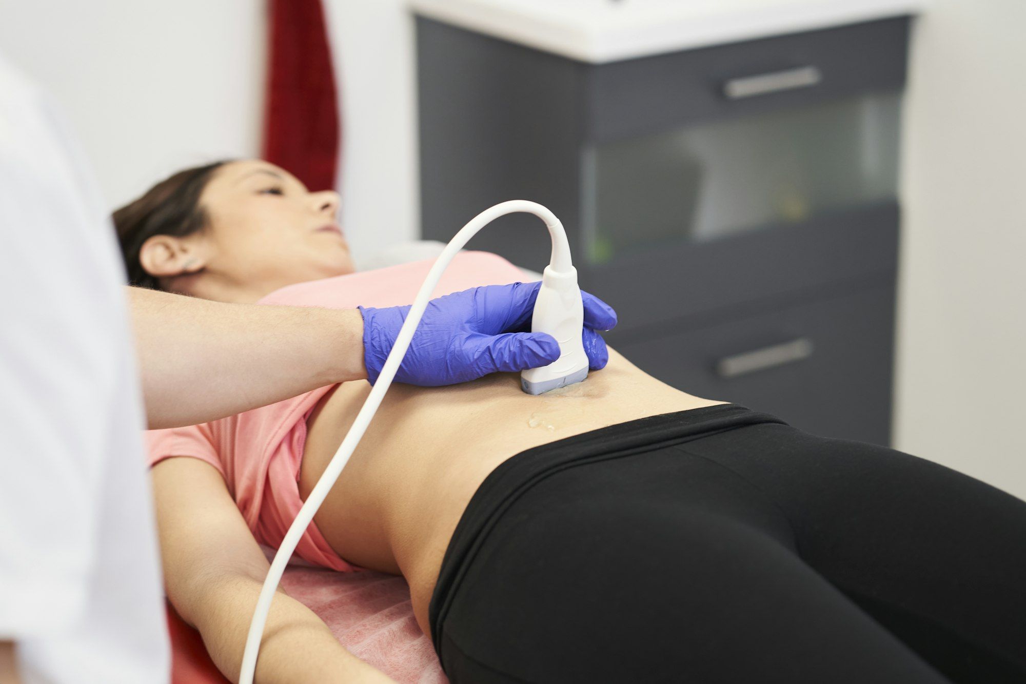 Pilates Canal Madrid Una persona recostada en una mesa de exploración recibe una ecografía abdominal. La persona que realiza la ecografía lleva guantes azules y sostiene el dispositivo sobre el abdomen descubierto del paciente. En el fondo se ve el equipo médico, que destaca los servicios de atención médica integral, incluidas las evaluaciones del suelo pélvico.