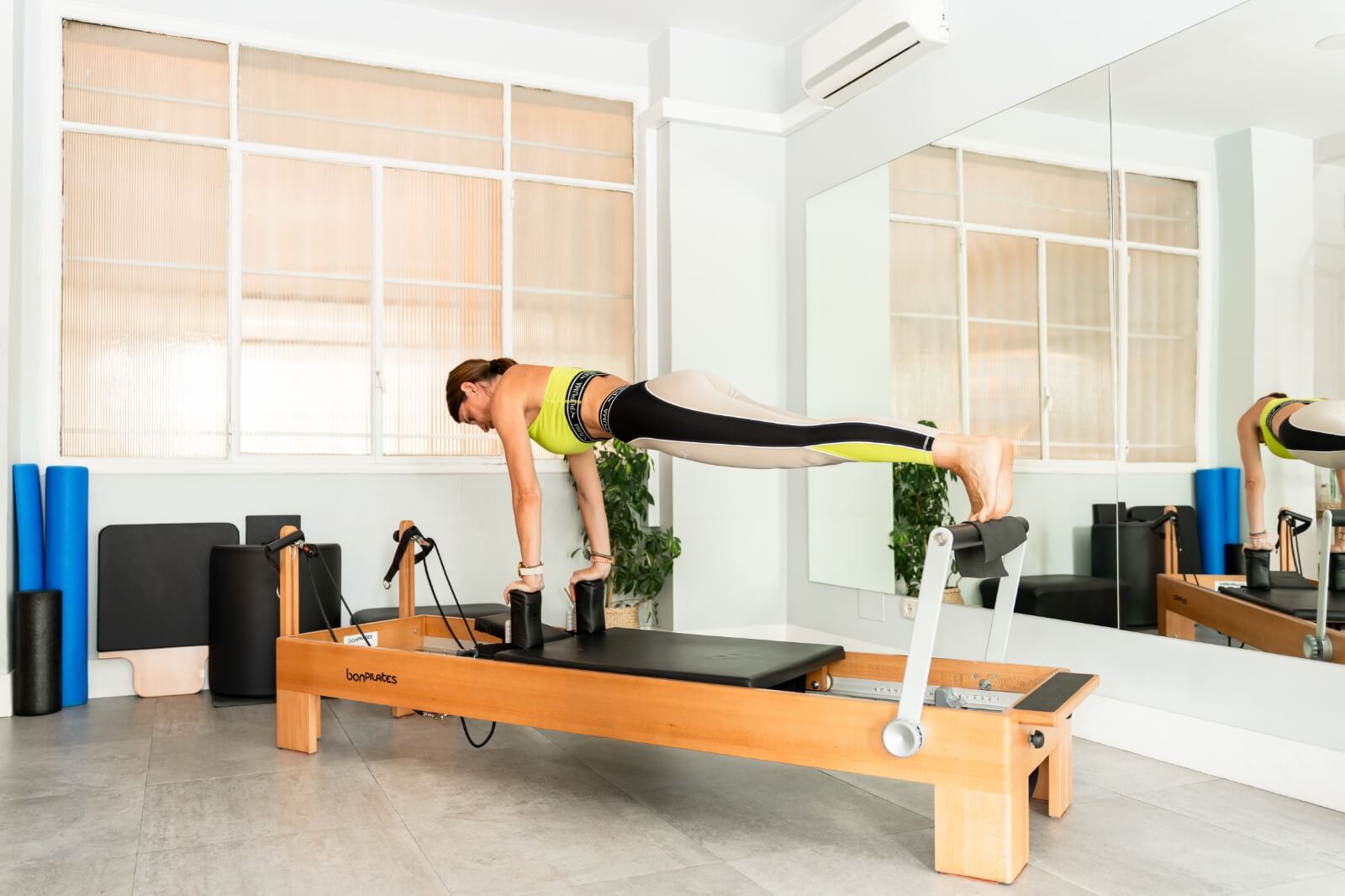 Pilates Canal Madrid En una sala luminosa con grandes ventanales y espejos, una persona con ropa deportiva muestra un ejercicio de plancha en un reformador de Pilates. En un ambiente de bienestar, los aparatos de fitness y las colchonetas enrolladas se disponen ordenadamente al fondo, evocando la esencia del bienestar madrileño.