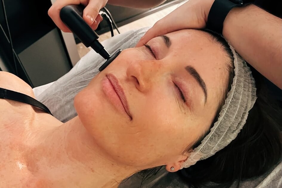 Pilates Canal Madrid Una persona yace tranquilamente en una camilla de spa en Madrid con los ojos cerrados, disfrutando de un tratamiento facial. Un dispositivo portátil se aplica suavemente sobre su mejilla, abrazando la terapia dermatofuncional. Con una diadema en su lugar, parece completamente relajada.