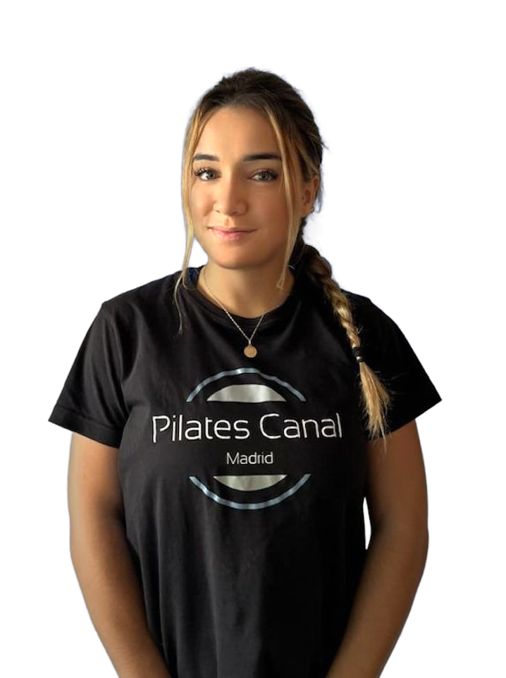 Pilates Canal Madrid Una mujer de pelo largo recogido en una trenza lleva una camiseta negra con la inscripción "Pilates Canal Madrid". Está de pie frente a un fondo liso y sonríe levemente mientras mira a la cámara, encarnando la esencia del Centro de bienestar en Madrid.