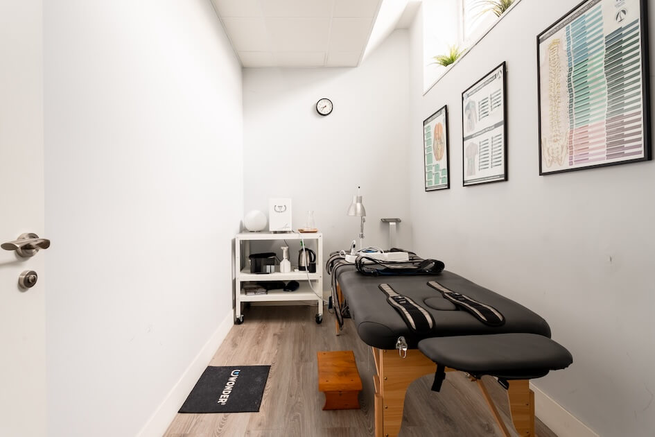 Pilates Canal Madrid Una pequeña y bien iluminada sala de tratamiento quiropráctico en Pilates Madrid cuenta con una mesa de tratamiento negra acolchada con correas, un pequeño taburete de madera y un carrito móvil blanco con suministros. Las paredes muestran historias clínicas mientras una puerta está ligeramente abierta en un lado.