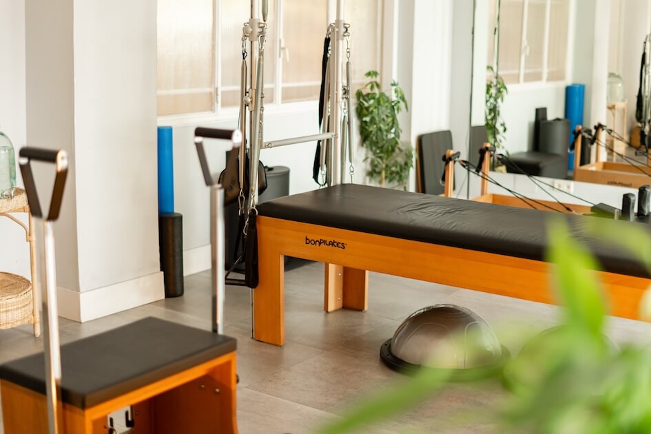 Pilates Canal Madrid Un estudio de Pilates bien iluminado en Madrid que cuenta con una máquina reformadora de la marca "BodyPilates" y otros equipos de Pilates. La sala tiene grandes ventanales, algunas plantas y espejos, lo que crea un espacio de ejercicio limpio y sereno.