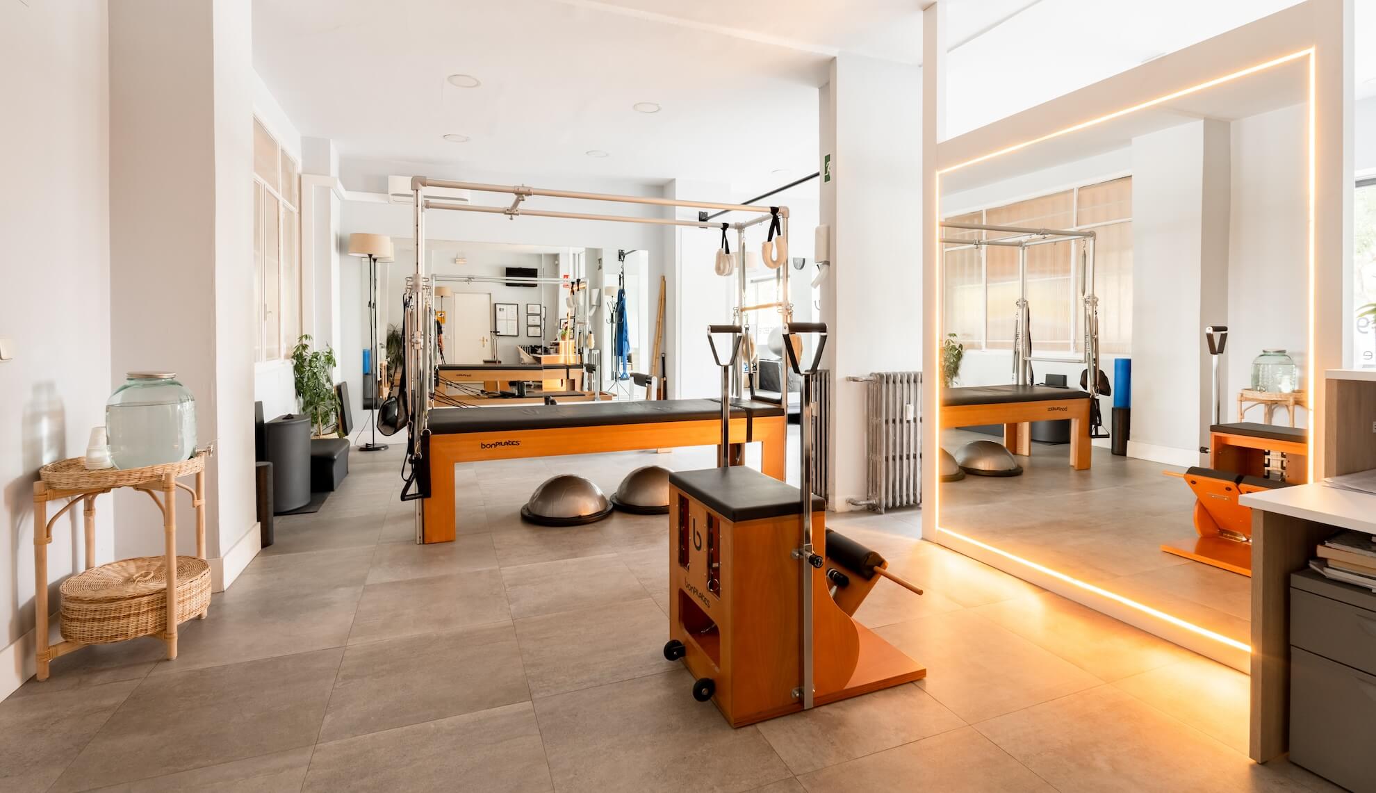 Pilates Canal Madrid Un estudio de pilates moderno y bien iluminado en Madrid que cuenta con varios equipos de pilates, incluidos reformers, una silla de ejercicios y domos de equilibrio. El estudio está decorado con paredes de colores claros, una gran pared con espejos e iluminación integrada y plantas verdes que crean un ambiente fresco.