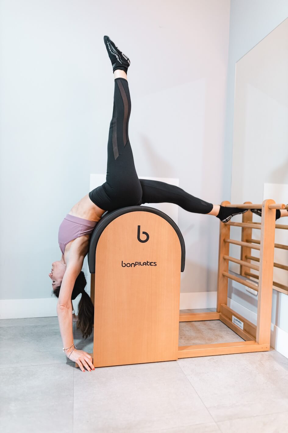Pilates Canal Madrid Una persona que lleva un sujetador deportivo morado y unas mallas negras realiza un ejercicio avanzado invertido en un barril de step de Bonpilates. Los pies de la persona se elevan sobre una escalera de madera, mientras que su torso se arquea hacia atrás con las manos en el suelo, demostrando flexibilidad y fuerza: una muestra inspiradora de los servicios de fitness.