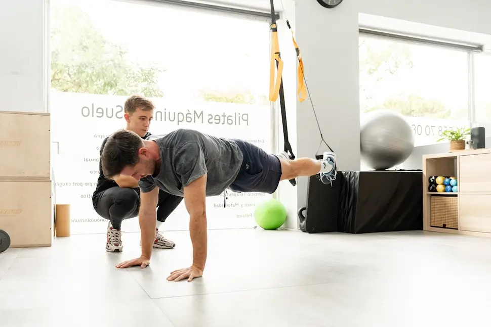 Pilates Canal Madrid Un hombre realiza flexiones suspendidas usando bandas de resistencia como parte de un ejercicio terapéutico mientras un entrenador se arrodilla cerca, observando su forma en un gimnasio moderno y luminoso con equipos de ejercicio en el fondo.