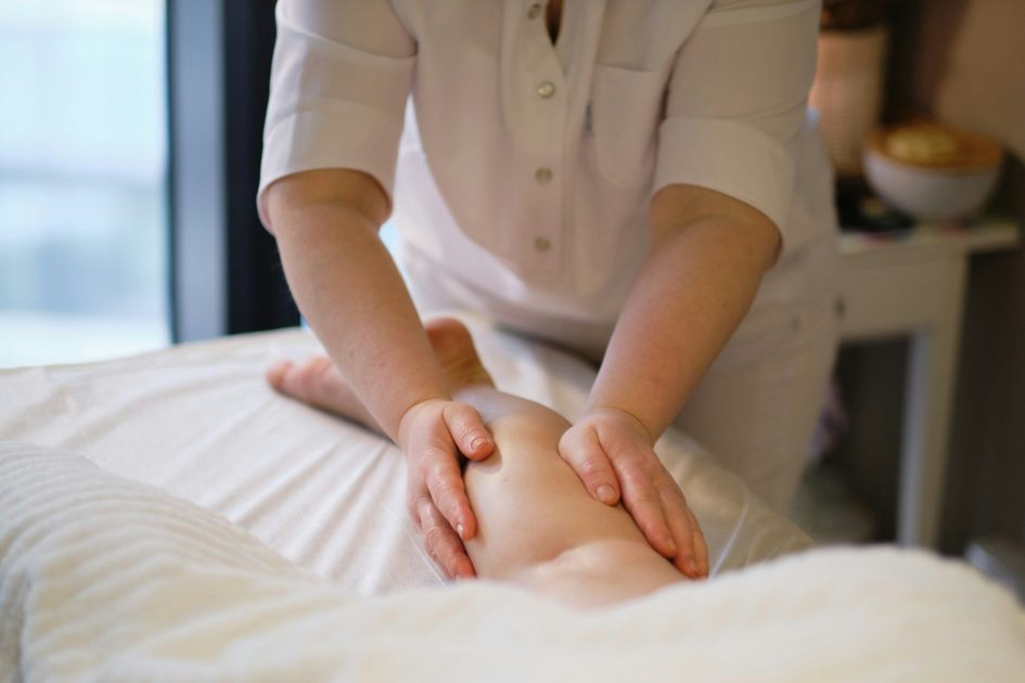 Pilates Canal Madrid Una persona vestida de blanco está dando un masaje, utilizando ambas manos para aplicar presión sobre la pierna de una persona tumbada boca abajo sobre una mesa de masajes cubierta con una sábana blanca. El entorno parece ser una sala de masajes o terapia profesional especializada en drenaje linfático manual fisioterapia madrid.