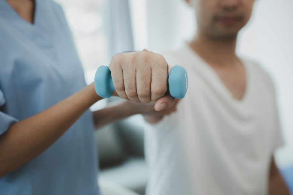 Pilates Canal Madrid Una persona con camisa blanca sostiene una pequeña mancuerna azul, asistida por un profesional sanitario que viste un uniforme azul. El profesional sanitario sostiene el brazo de la persona durante una sesión de ejercicio o fisioterapia en Fisioterapia Madrid, especializada en el tratamiento del dolor crónico.