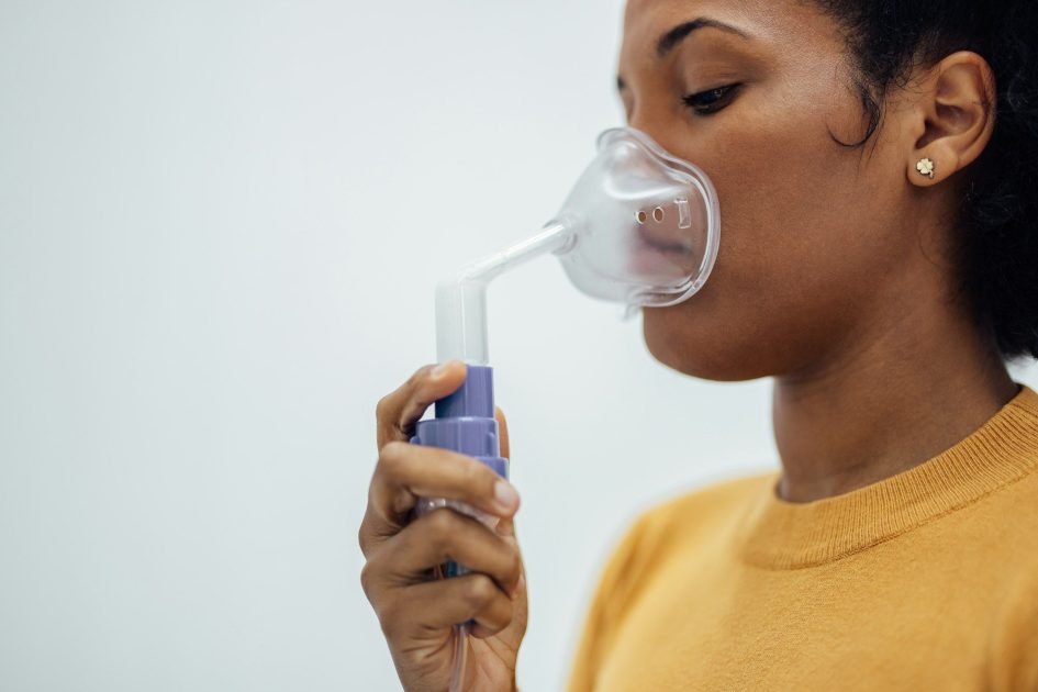 Pilates Canal Madrid Una persona con una camiseta amarilla utiliza una mascarilla nebulizadora para inhalar, lo que pone de relieve la importancia de la fisioterapia respiratoria en Madrid. La mascarilla transparente cubre la nariz y la boca, y el vapor que sale por el interior es visible. La persona sostiene el nebulizador en una mano e inhala el medicamento sobre un fondo blanco liso.