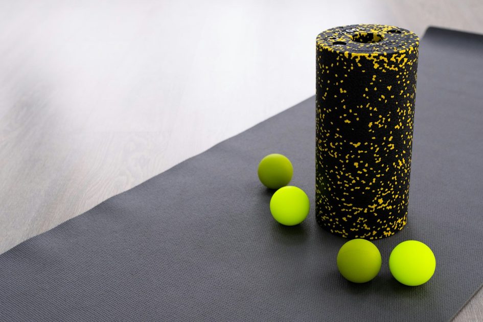 Pilates Canal Madrid Un rodillo de espuma negro con motas amarillas se encuentra en posición vertical sobre una colchoneta de ejercicios gris, junto a cuatro pelotas de masaje verdes. Este equipo de liberación miofascial está dispuesto sobre un suelo de madera clara, perfecto para rutinas de fisioterapia en Madrid.