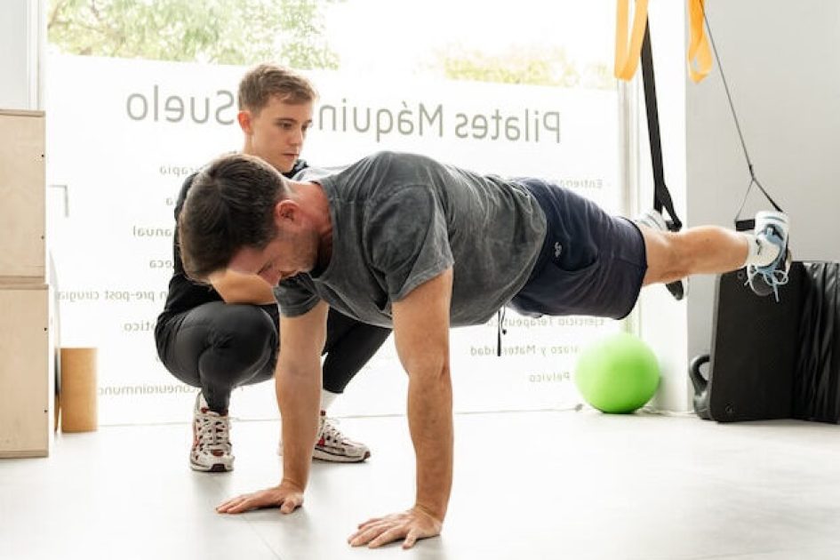 Pilates Canal Madrid Un hombre realiza ejercicios de plancha suspendida con los pies sujetos con correas TRX mientras un entrenador se arrodilla cerca y le ofrece orientación en un estudio de pilates moderno y luminoso en Madrid. Al fondo se ven varios aparatos de fitness como balones de estabilidad, pesas y cajas.