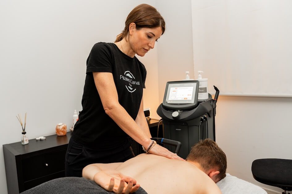 Pilates Canal Madrid Una masajista con camiseta negra le da un masaje de espalda a un cliente tumbado boca abajo en una camilla de masajes en un spa o sala de terapias. En un armario negro cercano hay varios productos para el cuidado de la piel y masajes. Junto a la camilla hay una máquina de masajes, lo que crea un ambiente similar al de los estudios de pilates de Madrid.