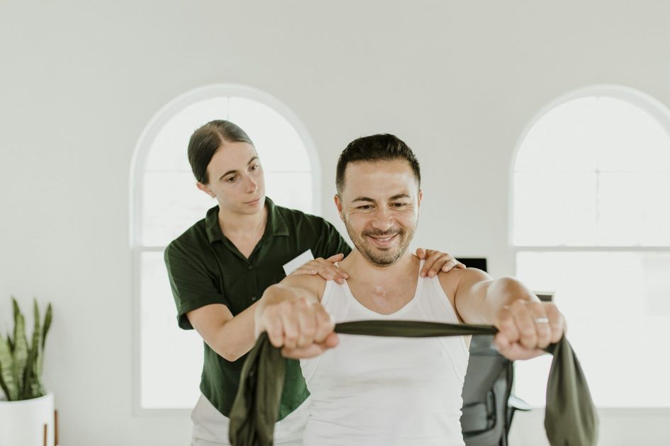 Pilates Canal Madrid Un hombre con camiseta blanca sin mangas utiliza una banda elástica, mientras una mujer con camiseta verde le ayuda colocando las manos sobre los hombros. Están recibiendo rehabilitación fisioterapia Madrid en una habitación luminosa con ventanas arqueadas al fondo y una planta en el lado izquierdo.