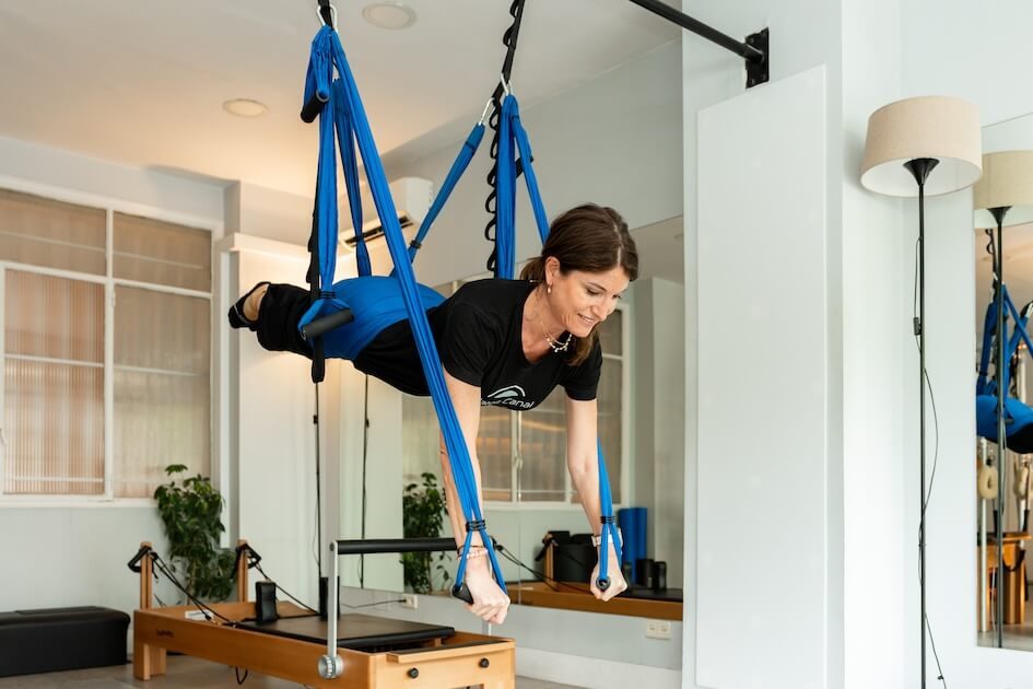 Pilates Canal Madrid Una persona practica yoga aéreo con gracia, suspendida en hamacas azules en un estudio tranquilo. Colocada horizontalmente y mirando hacia abajo con los brazos extendidos, está rodeada de paredes blancas, un espejo y máquinas de pilates que complementan sutilmente la escena.